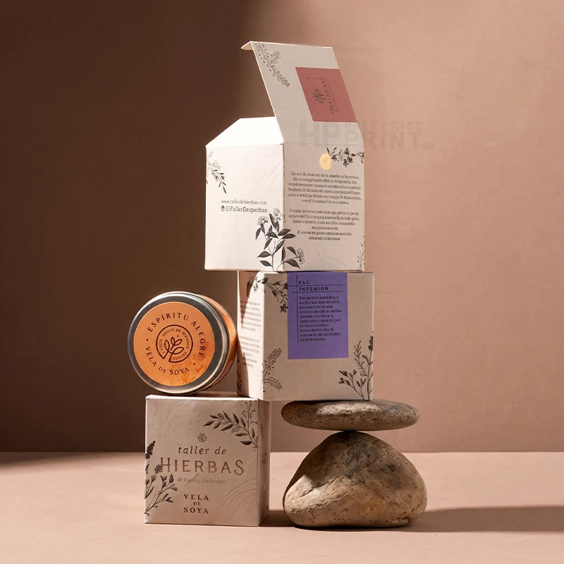 A styled product arrangement featuring a small round tin labeled 'A Spirit of Yucatan' and 'Taz de Miel' alongside two decorative tuck top boxes with botanical illustrations and a purple label, set against a warm brown background with stacked stones.