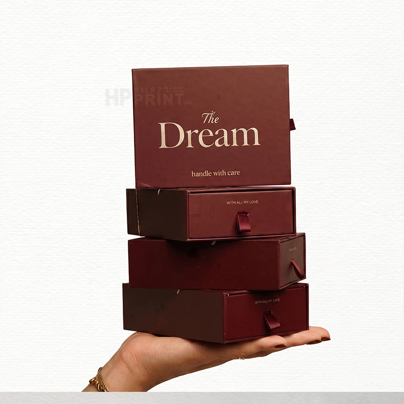 A stack of four dark burgundy gift drawer boxes with gold lettering, held in a person's hand against a light gray background. The top box reads 'The Dream' in gold serif text.