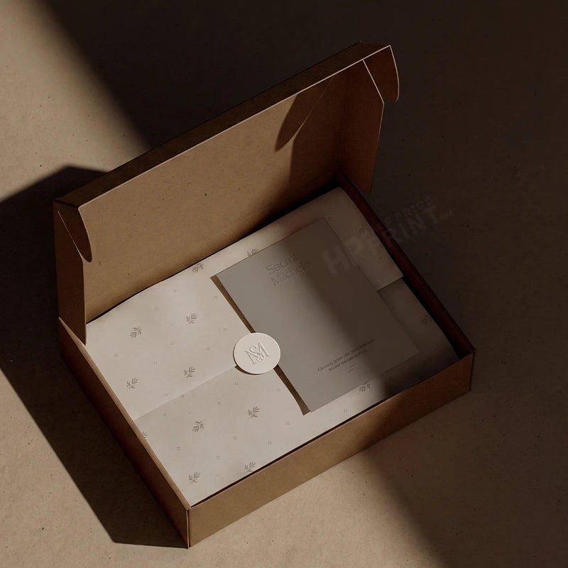 An open kraft paper gift box on a warm beige surface, lit by dramatic side lighting. Inside are white tissue papers with a small floral print and a gray card sealed with a round white sticker.