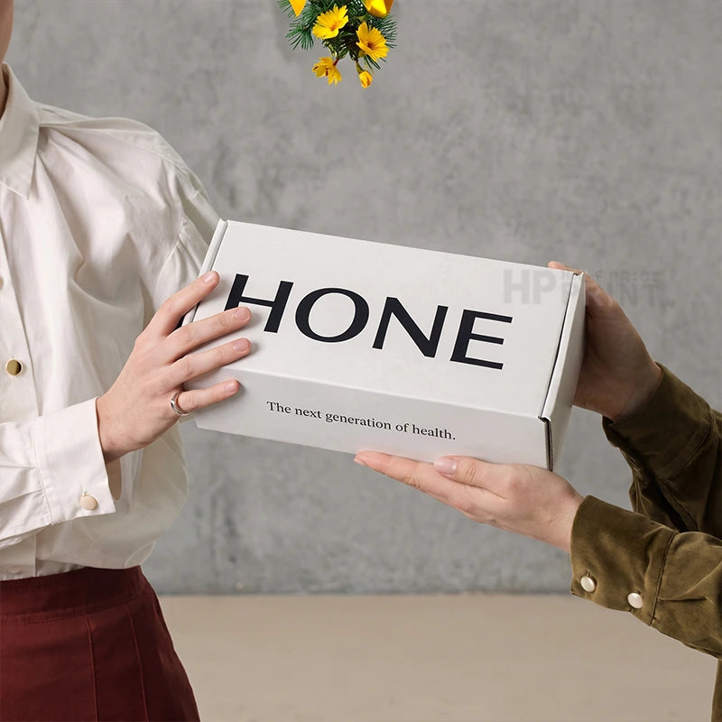 A person in a white shirt and burgundy skirt holds a white HONE mailer box with black lettering, while another person in an olive green top receives it, against a textured gray wall background.