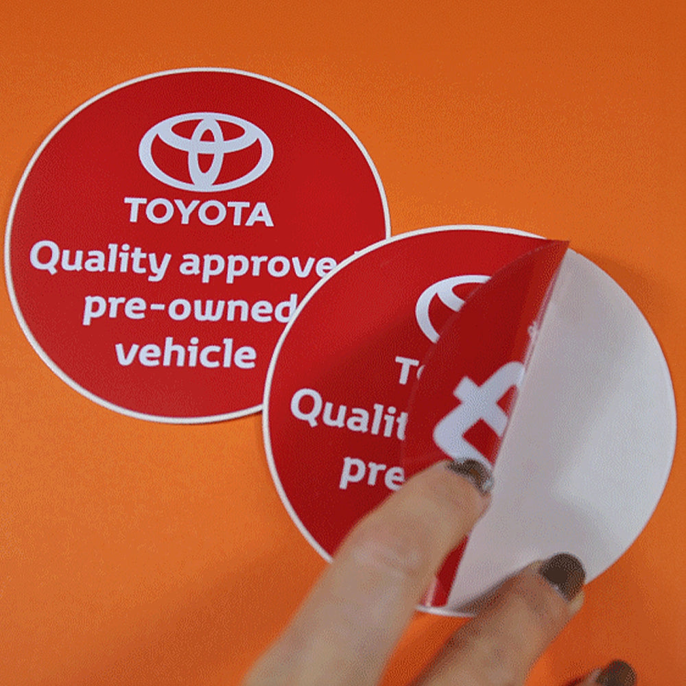A person peeling a red circular sticker with the Toyota logo and text 'Quality approved pre-owned vehicle' from a flat surface.