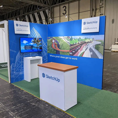 Trade show booth for SketchUp by cadsoftsolutions, featuring blue branded walls with architectural graphics and the slogan “Where great ideas get to work,” a mounted screen, and two white counters on green carpet inside an exhibition hall.