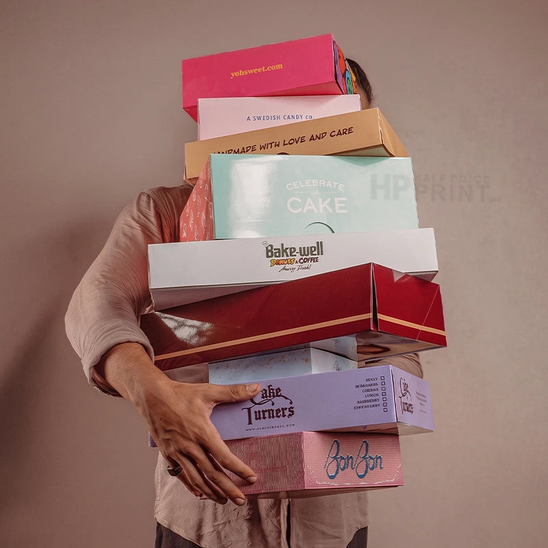 A person in a beige shirt carries a tall stack of colorful bakery cardboard gift boxes, including boxes for Cake Warehouse, Bake Well, and Jon Jon, against a solid taupe background.