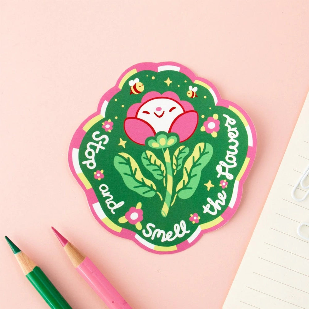 A die-cut sticker with a floral design and the text 'Stop and Smell the Flowers' on a pink background, accompanied by pencils and a notepad.