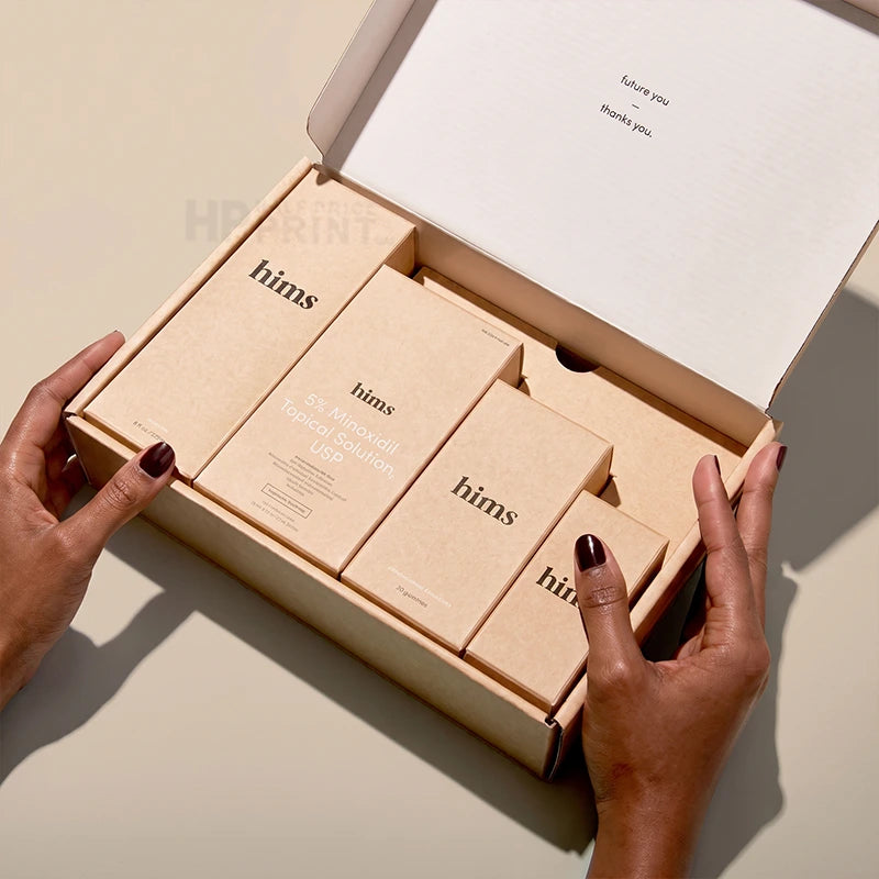 A pair of hands holding an open kraft paper gift mailer box containing several small beige product boxes printed with the brand name 'hims'. The scene is photographed on a warm beige surface with soft shadows.