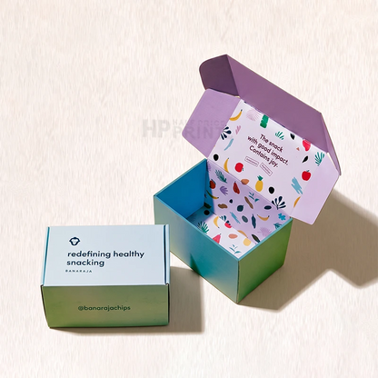 An open and closed product colored box set on a textured off-white background. The closed box is light blue on top with a green gradient base and reads 'redefining healthy snacking' with '@banarajachips'.