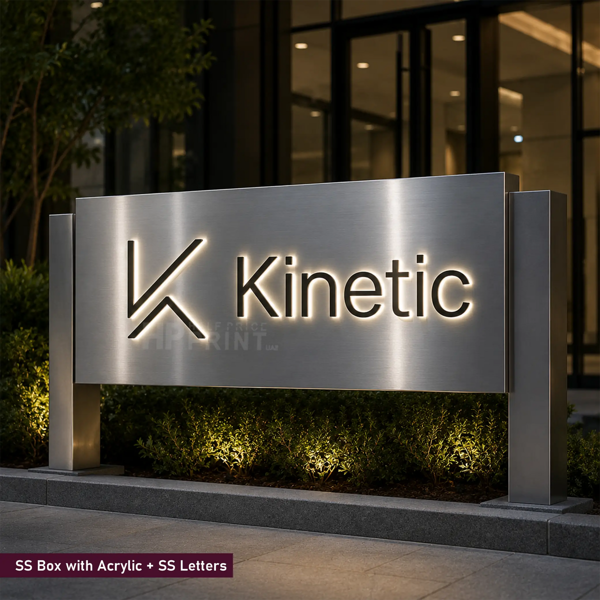 A large brushed stainless steel sign with the Kinetic logo and name illuminated in the background, installed outdoors in front of a modern building at night with landscaping and ground lights.