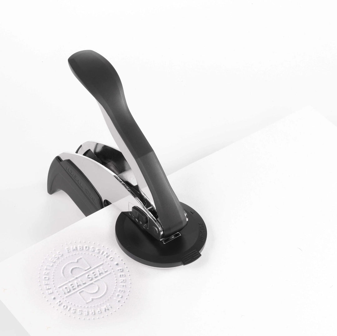 Handheld black and silver embossing stamp pressing a raised circular seal into white paper on a clean white background