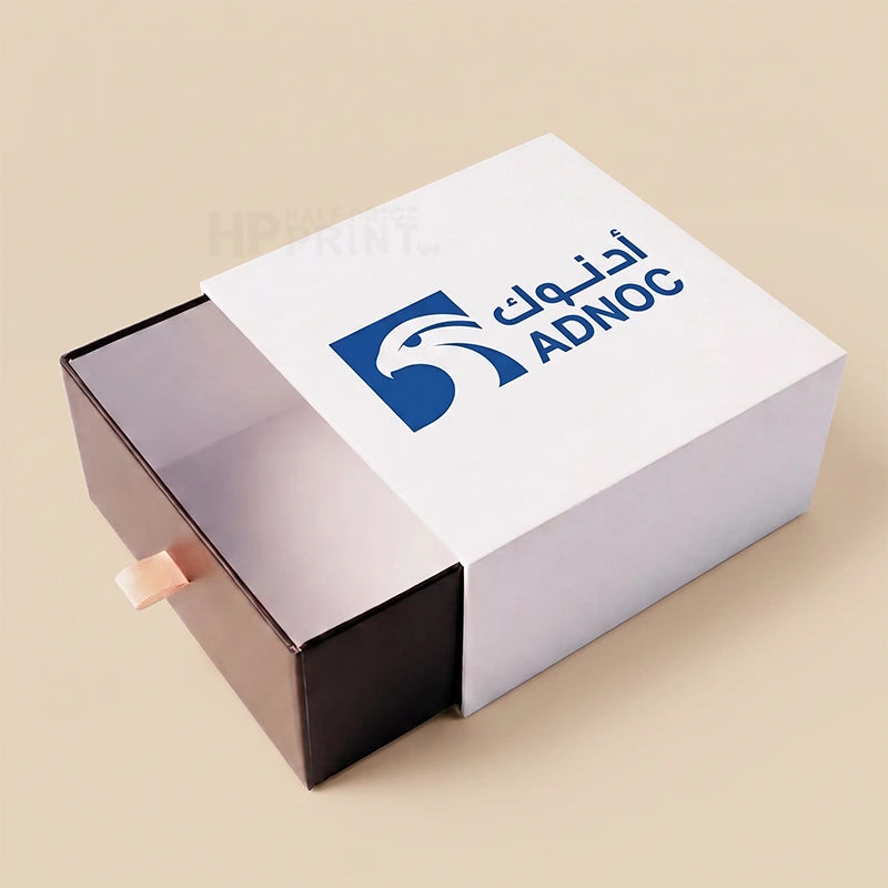 A white and black pull-out sliding box with a blue ADNOC logo printed on the top lid, set against a solid beige background.