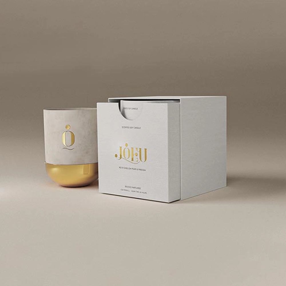 Minimalist product display of a luxury candle in a white and gold container beside a matching white presentation box with gold lettering, set against a neutral beige background.
