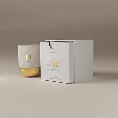 Minimalist product display of a luxury candle in a white and gold container beside a matching white presentation box with gold lettering, set against a neutral beige background.